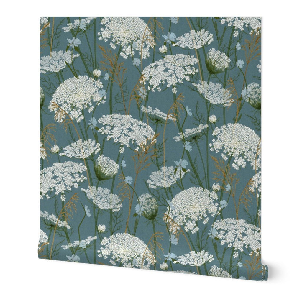 A roll of wallpaper featuring a repeating botanical pattern of white wildflowers on a muted blue-green background, shown against a plain white studio backdrop.