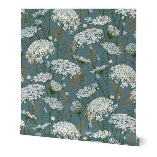A roll of wallpaper featuring a repeating botanical pattern of white wildflowers on a muted blue-green background, shown against a plain white studio backdrop.