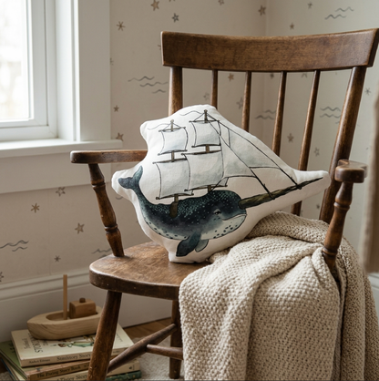 A decorative throw pillow a painted blue narwhal and sailing ship design, resting on a wooden chair in a softly lit room with wallpaper, a blanket, and a small toy boat.