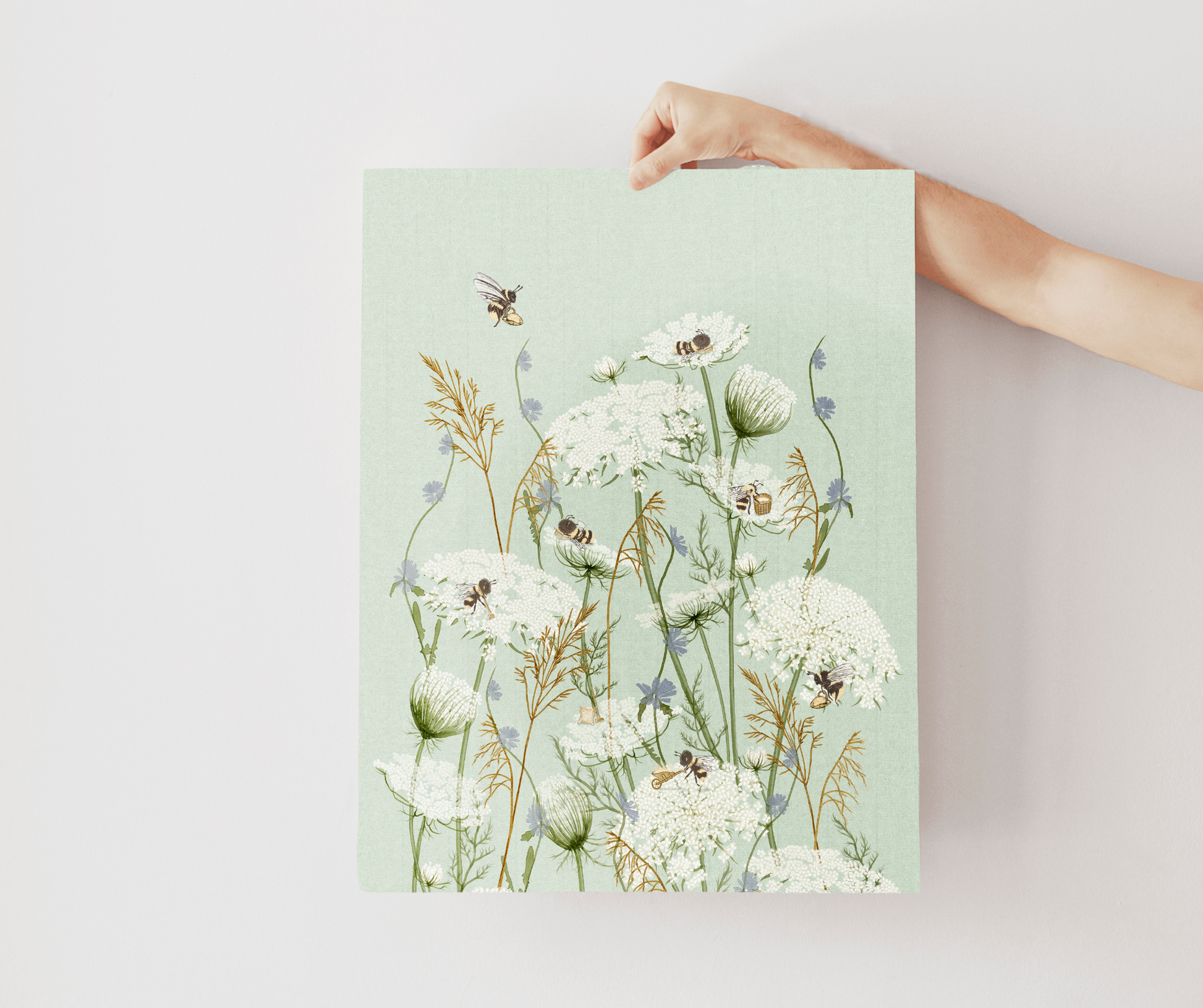 A hand holds a vertical art print featuring a botanical illustration of white wildflowers, bees, and dried grasses on a pale mint green background, set against a plain light gray wall.