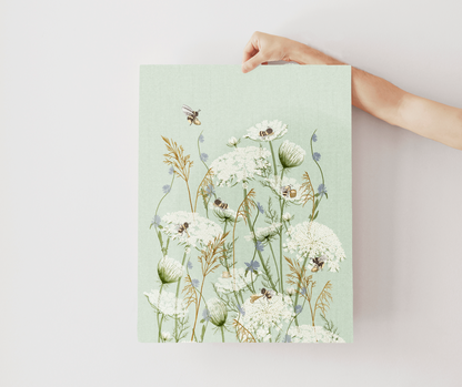 A hand holds a vertical art print featuring a botanical illustration of white wildflowers, bees, and dried grasses on a pale mint green background, set against a plain light gray wall.