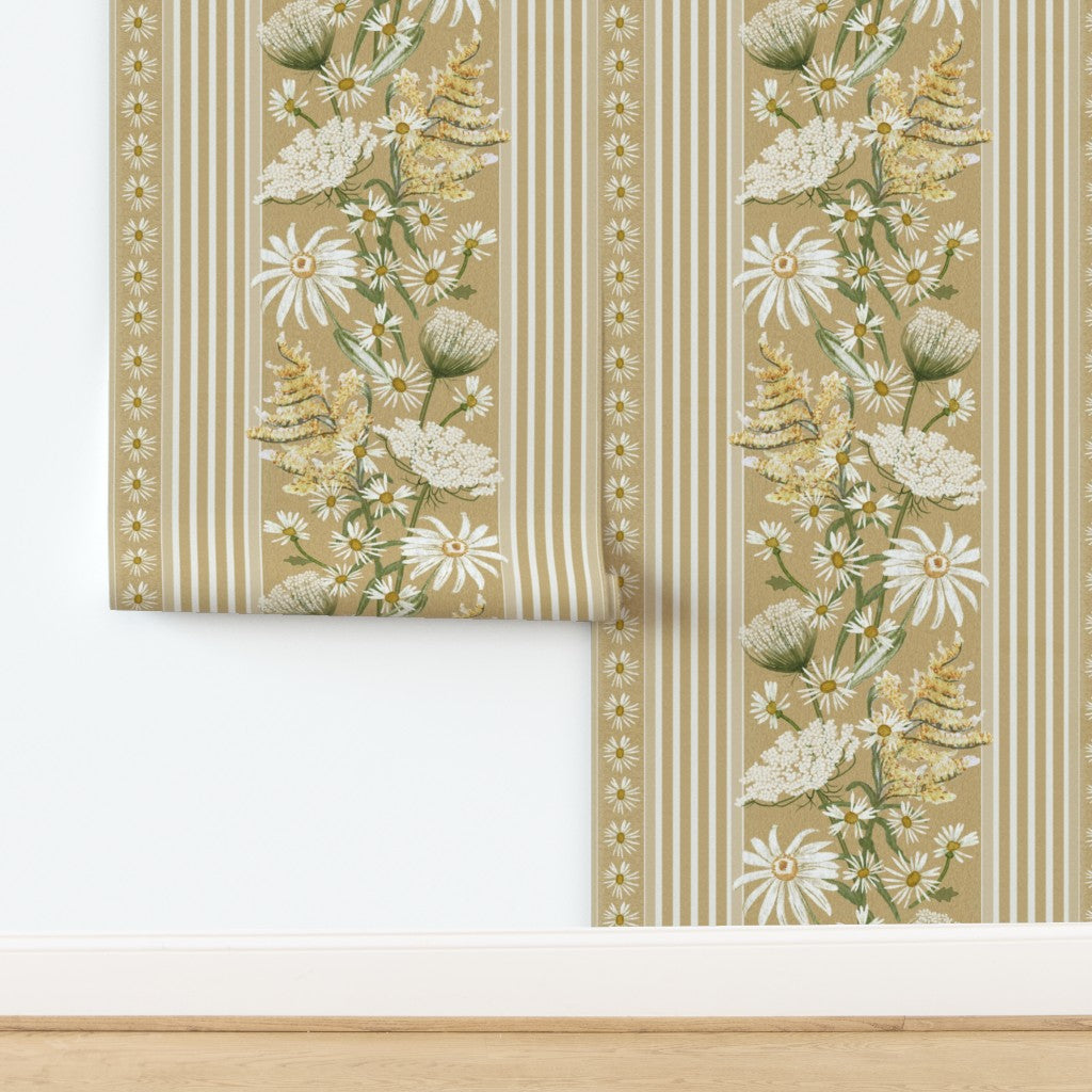 A roll of wallpaper partially unrolled on a white wall above a light wood floor, featuring a botanical floral pattern with daisies, wildflowers, and striped borders on a warm beige background.