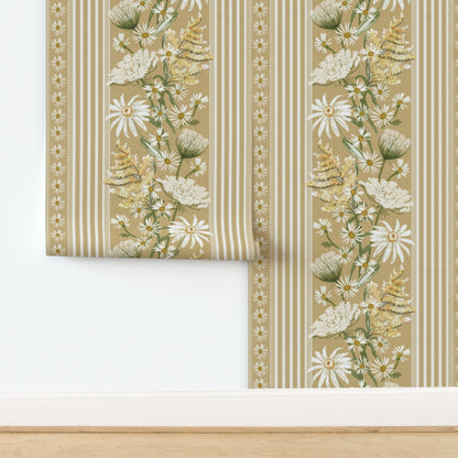 A roll of wallpaper partially unrolled on a white wall above a light wood floor, featuring a botanical floral pattern with daisies, wildflowers, and striped borders on a warm beige background.