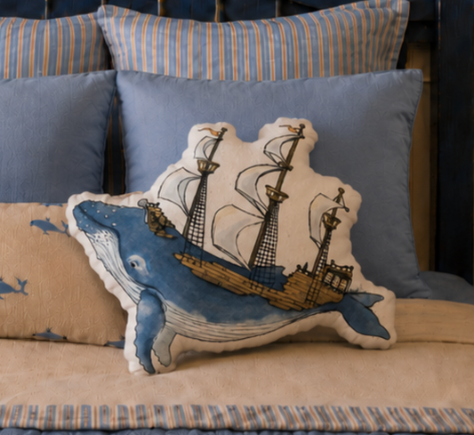 A whale-shaped throw pillow featuring a blue whale and a sailing ship, styled on a bed with blue and striped pillows.