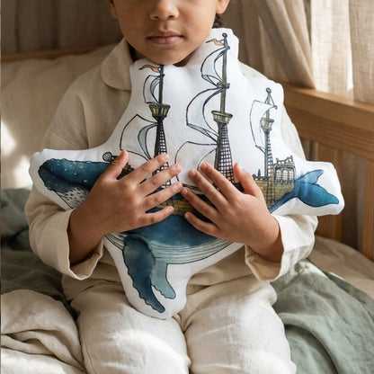 A child wearing a beige linen outfit holds a white plush pillow featuring a blue whale and pirate ship illustration.
