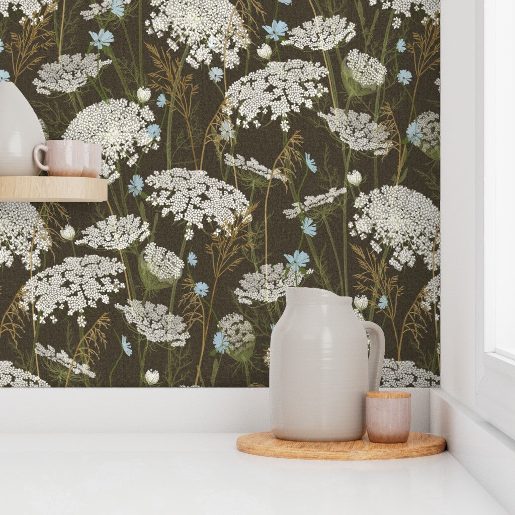 A styled interior scene showing a wall covered in dark brown floral wallpaper with white, blue, and tan wildflower motifs. A white ceramic pitcher and small cup sit on a round wooden tray on a white countertop, with a shelf and vase partially visible on the left.