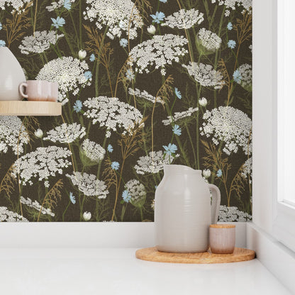 A styled interior scene showing a wall covered in dark brown floral wallpaper with white, blue, and tan wildflower motifs. A white ceramic pitcher and small cup sit on a round wooden tray on a white countertop, with a shelf and vase partially visible on the left.