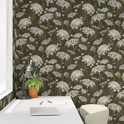 A room corner featuring dark brown wallpaper with a repeating white and light blue wildflower pattern, styled with a white desk, a potted green plant, and a cream-colored chair.
