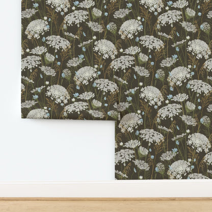 A roll of floral wallpaper with a dark brown background and a repeating pattern of white Queen Anne's lace flowers and small light blue wildflowers, shown installed on a white wall above a light wood floor.