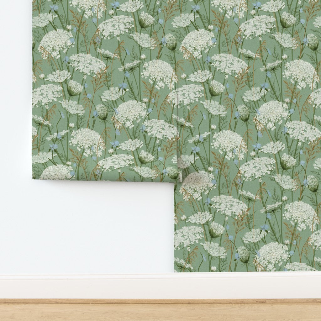 A folded piece of wallpaper with a repeating wildflower pattern in neutral green, white, and soft blue, displayed against a white wall above a light wood floor.