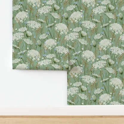 A folded piece of wallpaper with a repeating wildflower pattern in neutral green, white, and soft blue, displayed against a white wall above a light wood floor.