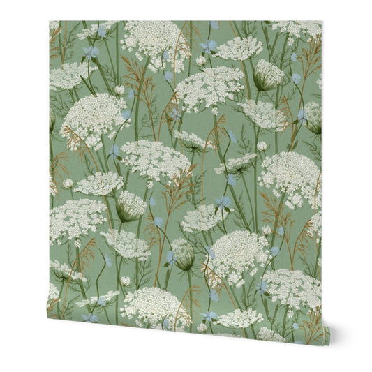 wallpaper featuring a repeating botanical pattern of white wildflowers, blue blossoms, and brown grasses on a muted gray green background, shown against a clean white studio backdrop.