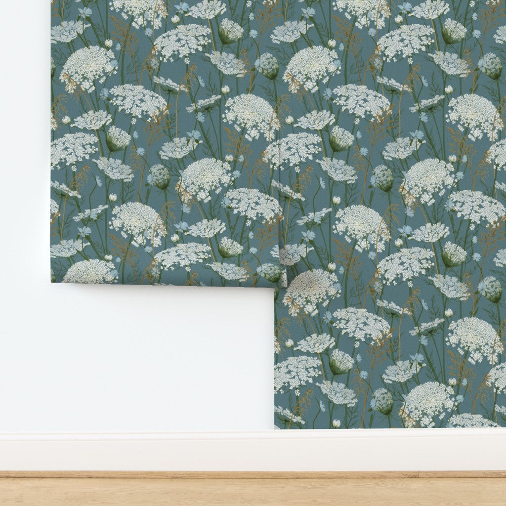 A folded piece of wallpaper  with a repeating white wildflower pattern on a muted blue-gray background, displayed against a white wall above a light wood floor.