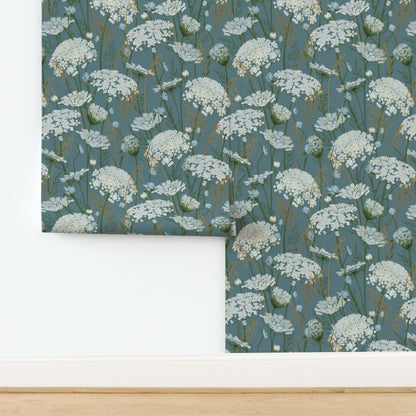 A folded piece of wallpaper  with a repeating white wildflower pattern on a muted blue-gray background, displayed against a white wall above a light wood floor.