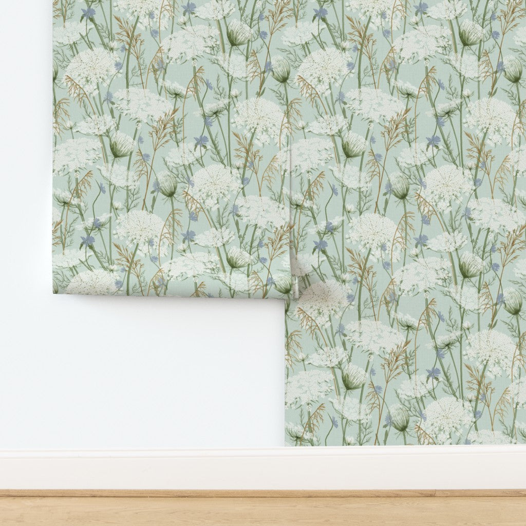 A roll of wallpaper with a botanical pattern of white flowers and green stems on a pale aqua background, shown partially unrolled on a white wall above a light wood floor.