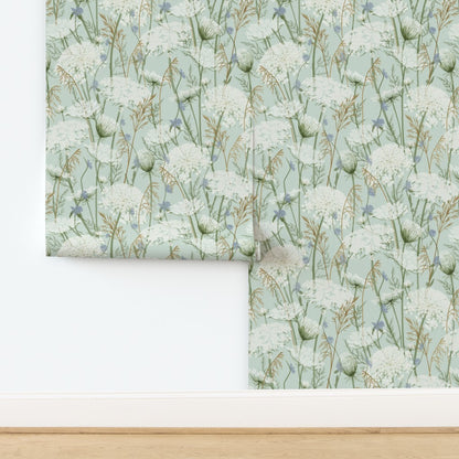 A roll of wallpaper with a botanical pattern of white flowers and green stems on a pale aqua background, shown partially unrolled on a white wall above a light wood floor.