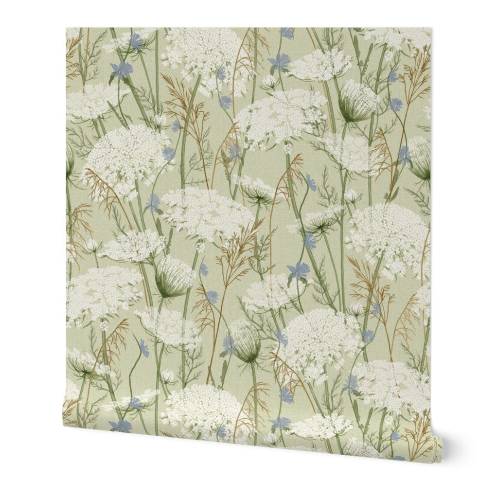 Wallpaper featuring a delicate botanical pattern of white wildflowers, blue blossoms, and green stems on a pale sage green background, shown against a plain white studio backdrop.