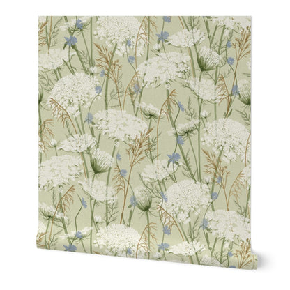 Wallpaper featuring a delicate botanical pattern of white wildflowers, blue blossoms, and green stems on a pale sage green background, shown against a plain white studio backdrop.