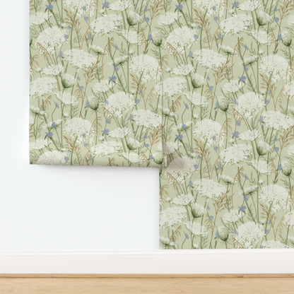 A folded piece of wallpaper with a delicate botanical pattern of white flowers and green stems on a pale sage green background, displayed against a white wall with a light wood floor visible at the bottom.